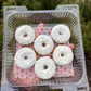 Plastic container with six white frosted donuts on a pink heart-patterned paper against a natural background.