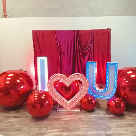 Luxury 'I ❤️ U' balloon installation with a red and blush balloon garland, floral accents, and oversized metallic spheres. A woman poses in front of the glowing setup.