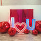 Luxury 'I ❤️ U' balloon installation with a red and blush balloon garland, floral accents, and oversized metallic spheres. A woman poses in front of the glowing setup.