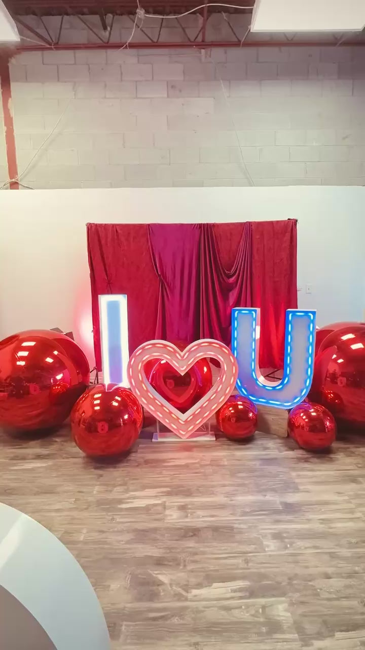 Luxury 'I ❤️ U' balloon installation with a red and blush balloon garland, floral accents, and oversized metallic spheres. A woman poses in front of the glowing setup.