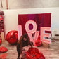 Romantic event decor featuring illuminated 'LOVE' letters, an elegant floral and balloon arrangement, and a red drapery backdrop. A woman sits in front, smiling at the camera.