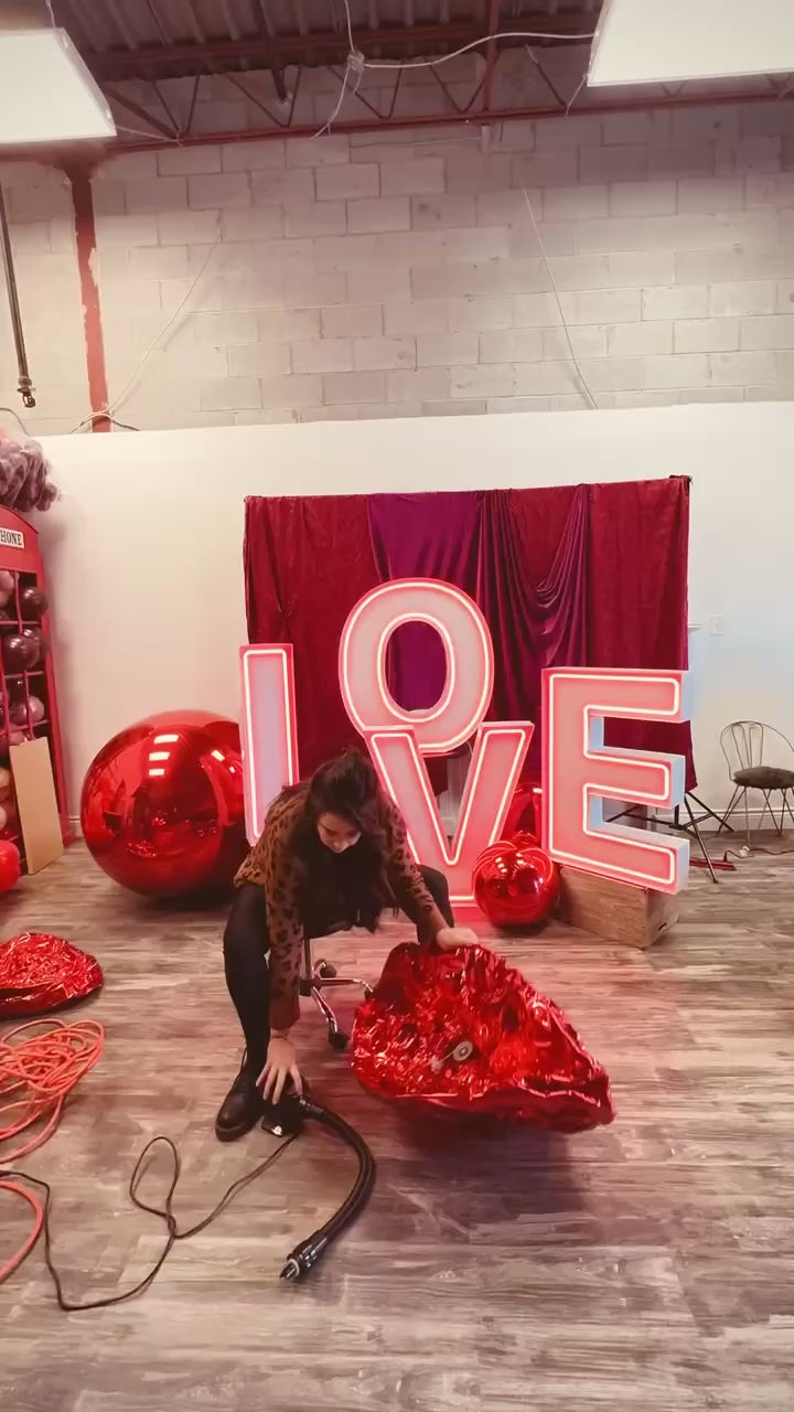 Romantic event decor featuring illuminated 'LOVE' letters, an elegant floral and balloon arrangement, and a red drapery backdrop. A woman sits in front, smiling at the camera.