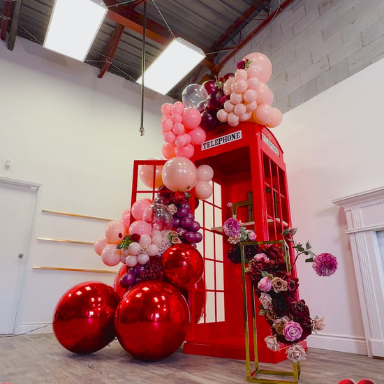 Luxury balloon decor with a red phone booth backdrop, blush and burgundy balloon garland, metallic spheres, and floral details. A woman poses playfully, showcasing the event setup