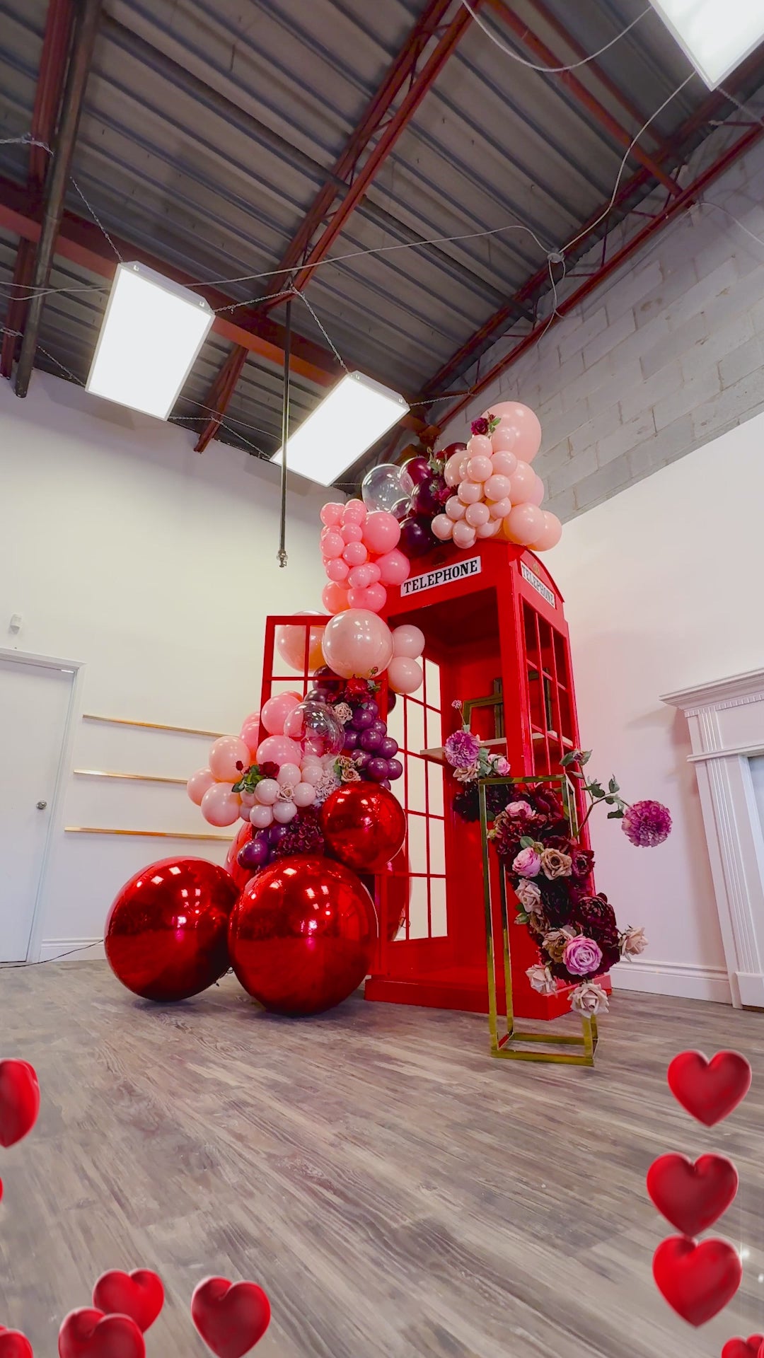 Luxury balloon decor with a red phone booth backdrop, blush and burgundy balloon garland, metallic spheres, and floral details. A woman poses playfully, showcasing the event setup