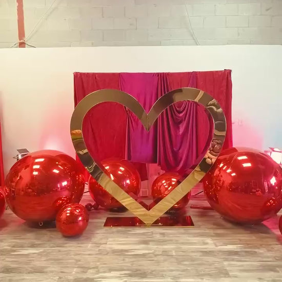 Valentine’s Day balloon setup with a gold heart frame, red balloon clusters, floral decor, and mirror balls. A woman smiles while standing in front of the display