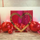 Valentine’s Day balloon setup with a gold heart frame, red balloon clusters, floral decor, and mirror balls. A woman smiles while standing in front of the display