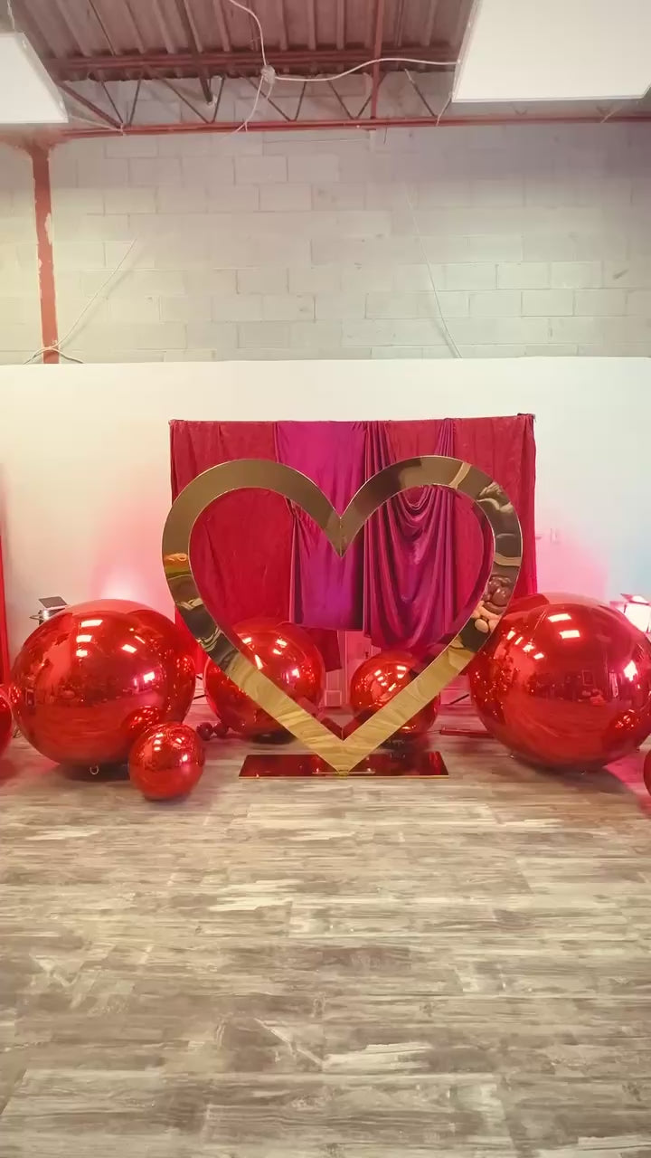 Valentine’s Day balloon setup with a gold heart frame, red balloon clusters, floral decor, and mirror balls. A woman smiles while standing in front of the display