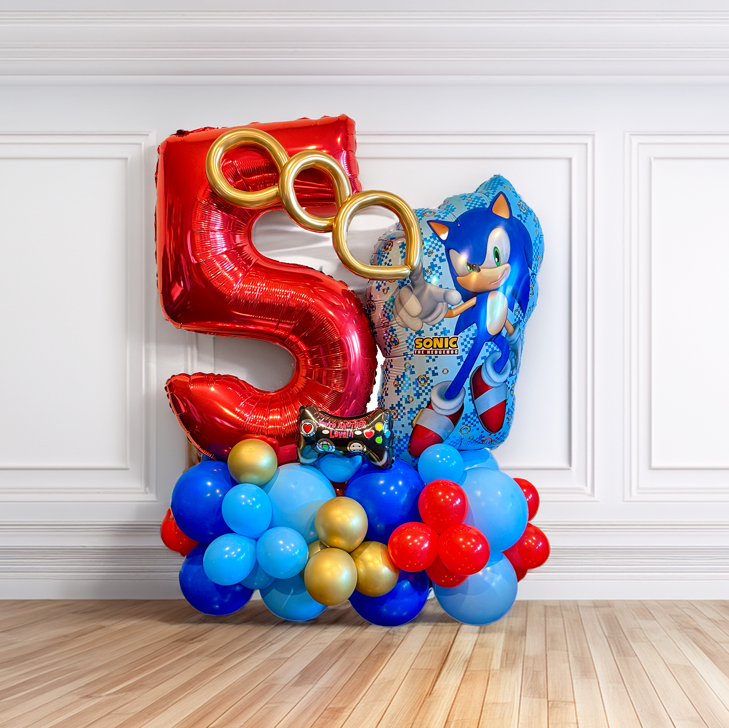 A balloon display featuring a large red number 5, Sonic the Hedgehog character balloon, gold balloon rings, and a variety of blue, red, and gold balloons arranged on a floor against a wall, representing a Sonic themed birthday celebration.